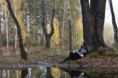 Woman resting under a tree by a reflective lake.